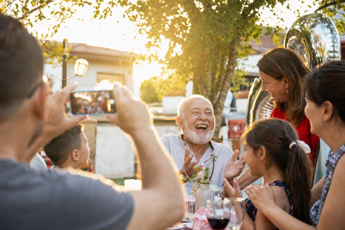 A family films a laughing grandfather with a smartphone at a backyard birthday party